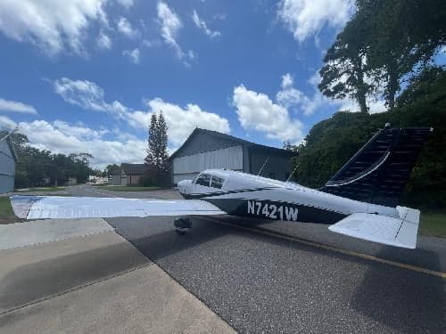 A rear-side view of the white and dark blue Piper Cherokee 180 aircraft, registration N7421W, parked on an asphalt surface with a hangar and trees in the background.