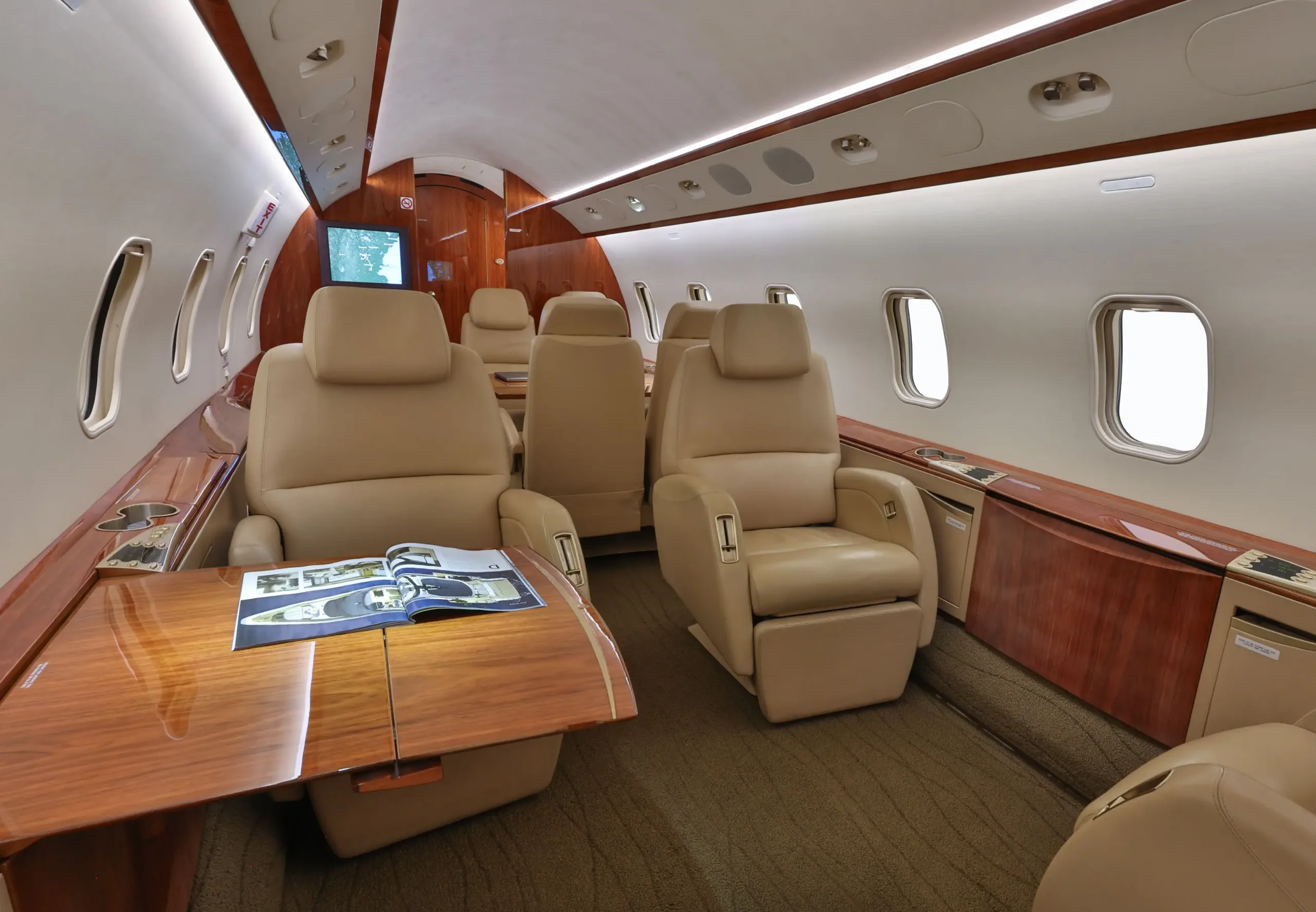 Another view of the aircraft cabin, showing beige leather seats, a polished wood table with a magazine, and a screen at the front.