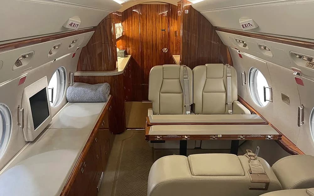 The aft section of a private jet cabin, featuring a side-facing divan that converts to a bed, a two-seat configuration with a table, and rich wood paneling.
