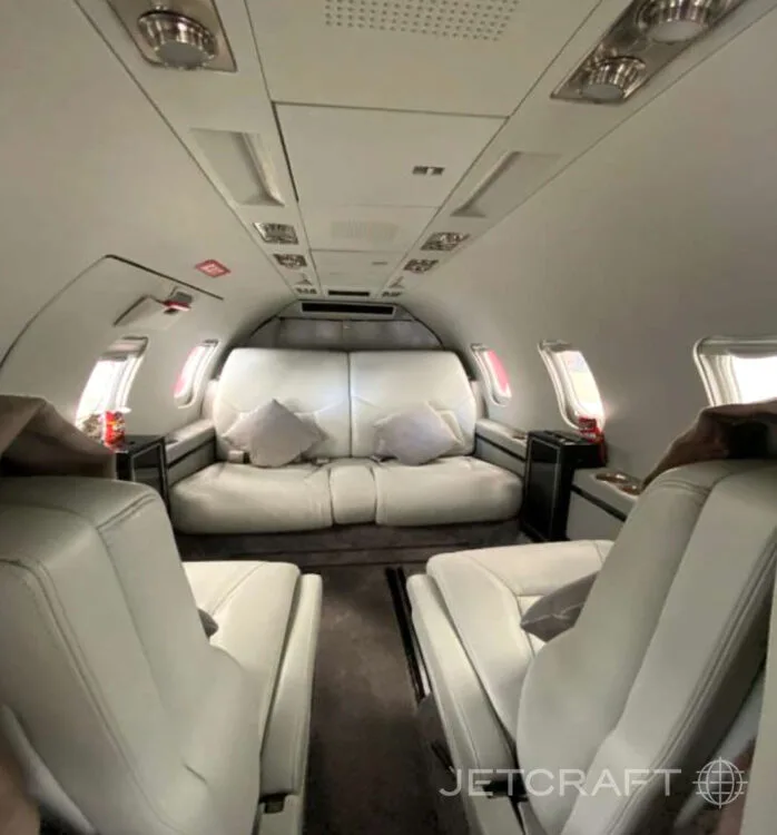 The interior cabin of a business jet, showing light gray leather seats and a matching sofa at the rear.