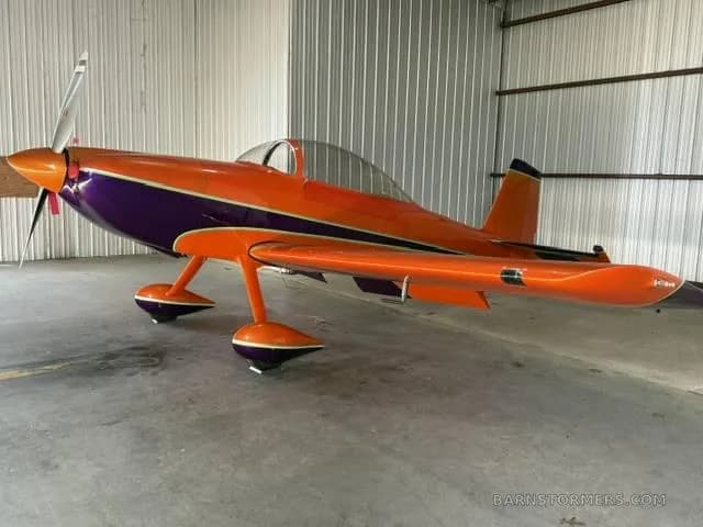 A three-quarter front view of an orange and purple single-engine aerobatic aircraft in a hangar.
