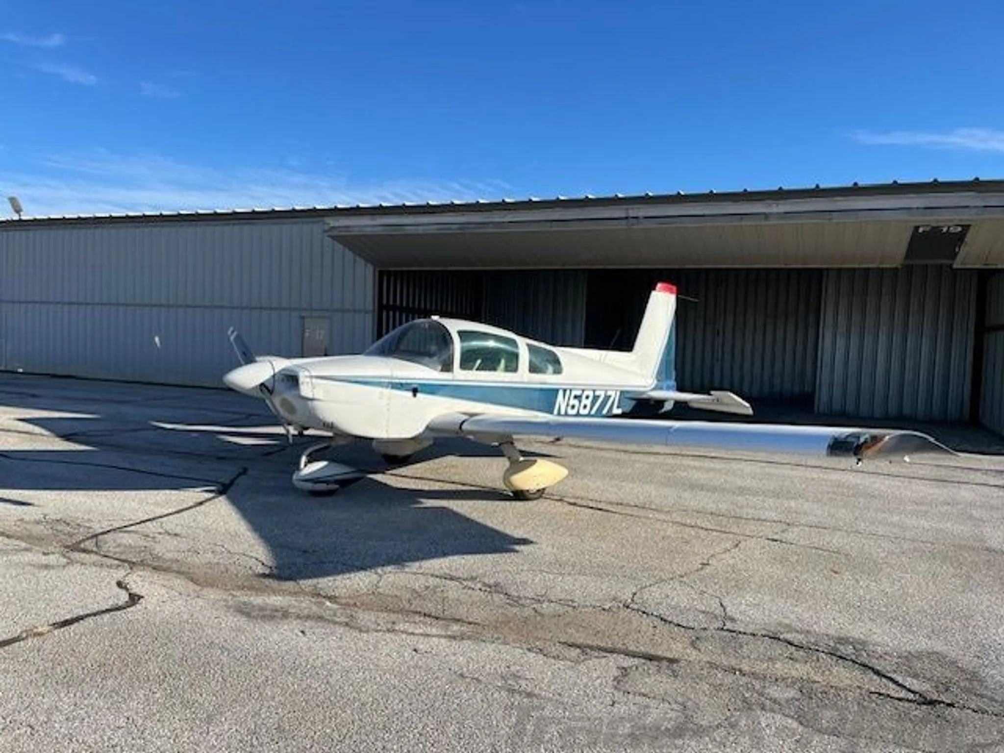A white and blue Grumman AA-5B Tiger aircraft (N5877L) parked on a tarmac with a hangar in the background.