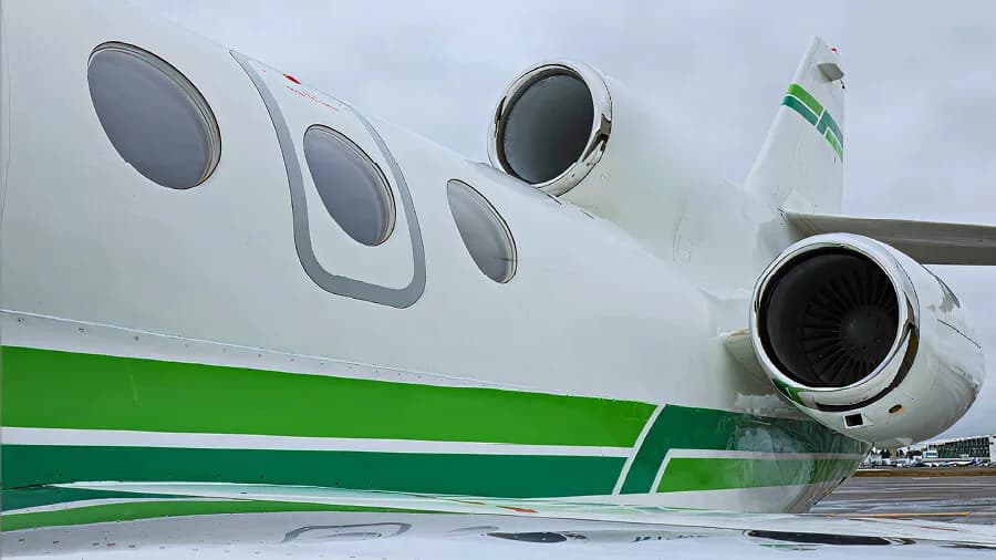 A close-up of the rear fuselage of a white business jet with green stripes, showing the engines and oval windows.