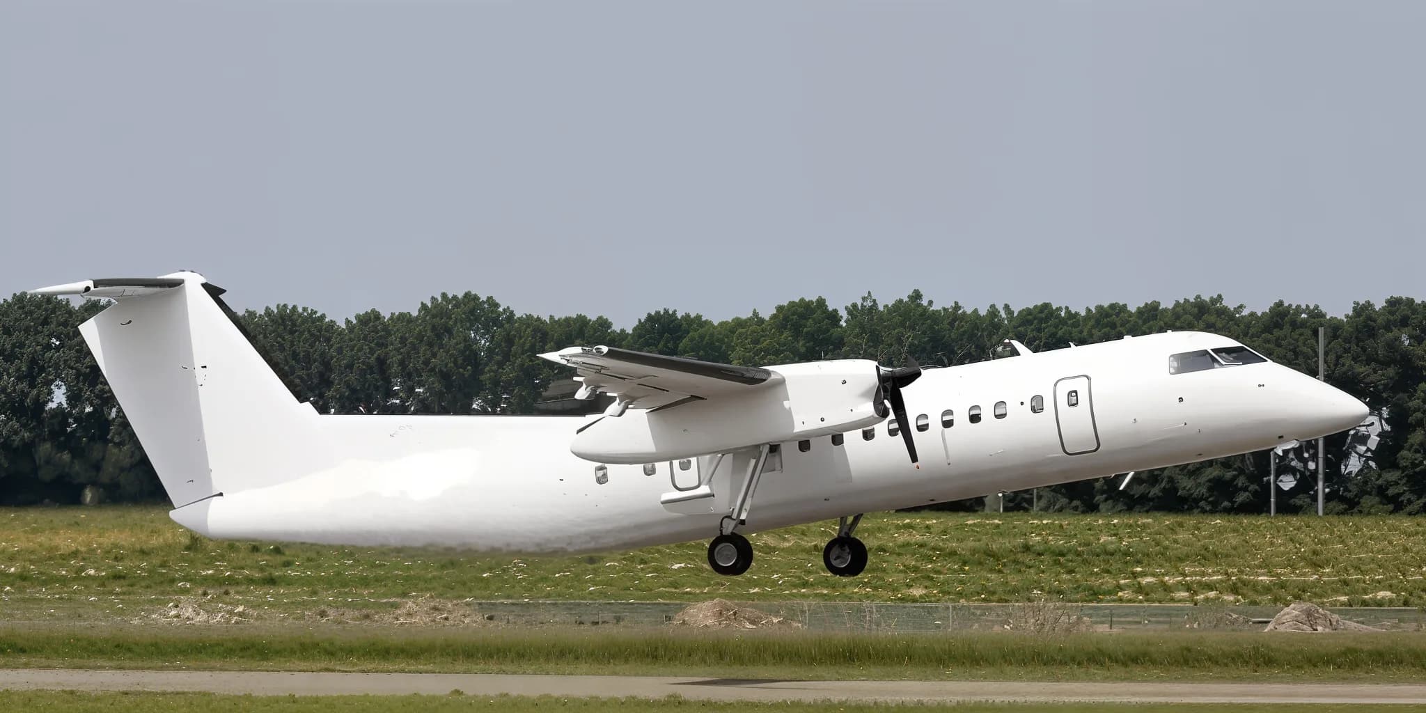 A white turboprop aircraft with its landing gear extended, just above a runway with trees in the background.
