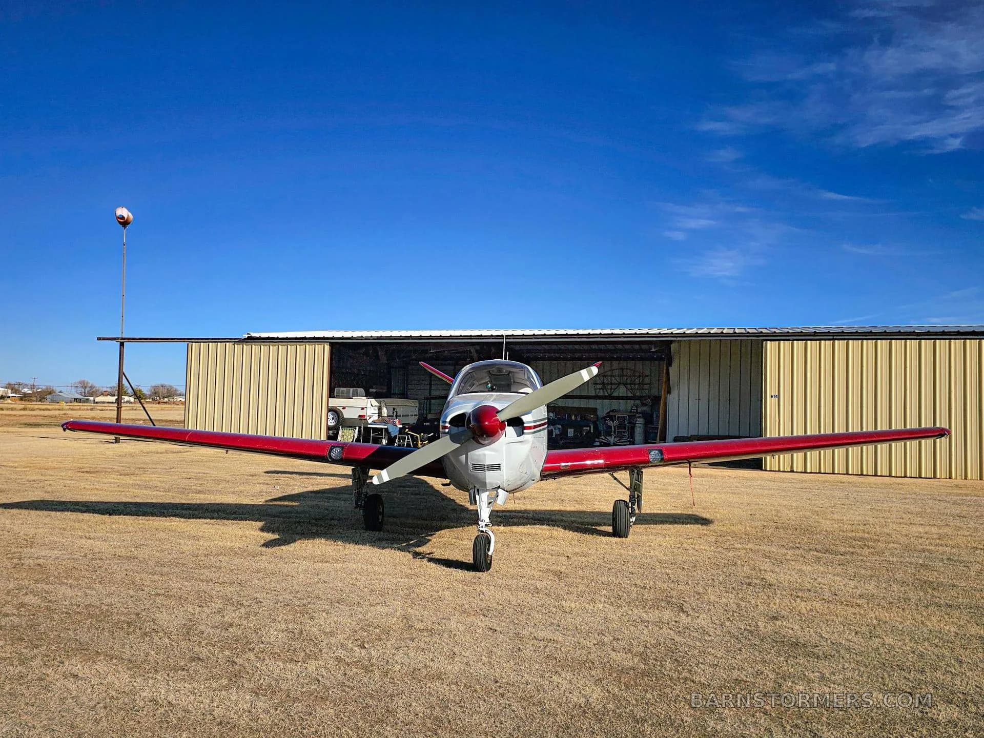 A white and red Mooney M20 series aircraft viewed from the front, parked on a grassy field in front of an open hangar.