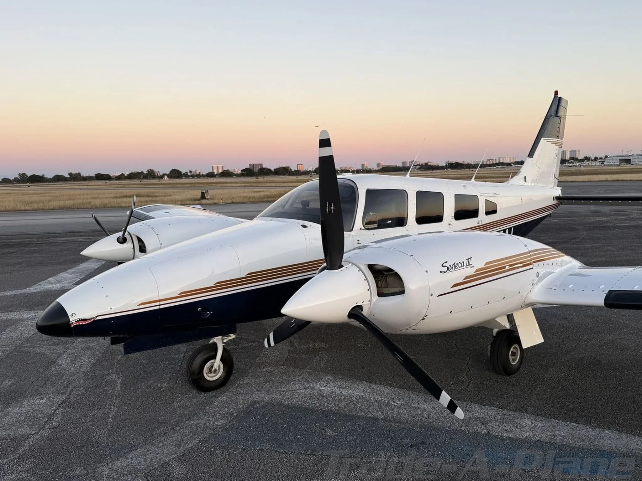 A Piper Seneca III twin-engine aircraft with white and blue paint scheme and gold stripes, parked on an airfield at sunset.