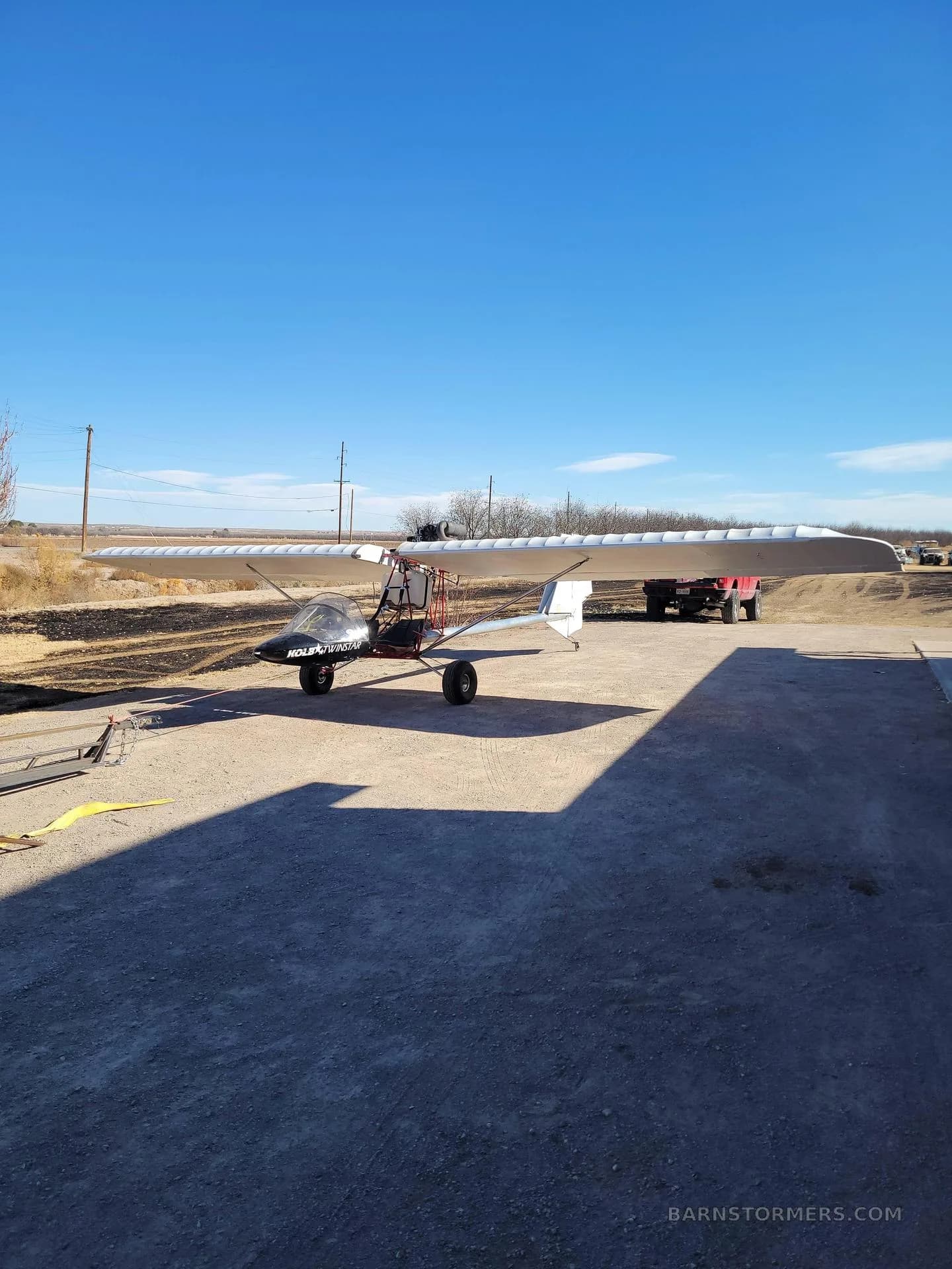 A black and white Volmer VJ-24W SunFun experimental aircraft is parked on a gravel lot under a clear blue sky.