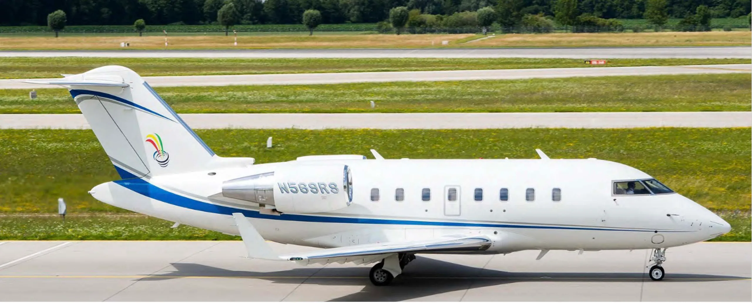 A white Bombardier Challenger business jet with blue and grey stripes and a tail logo, parked on a tarmac.