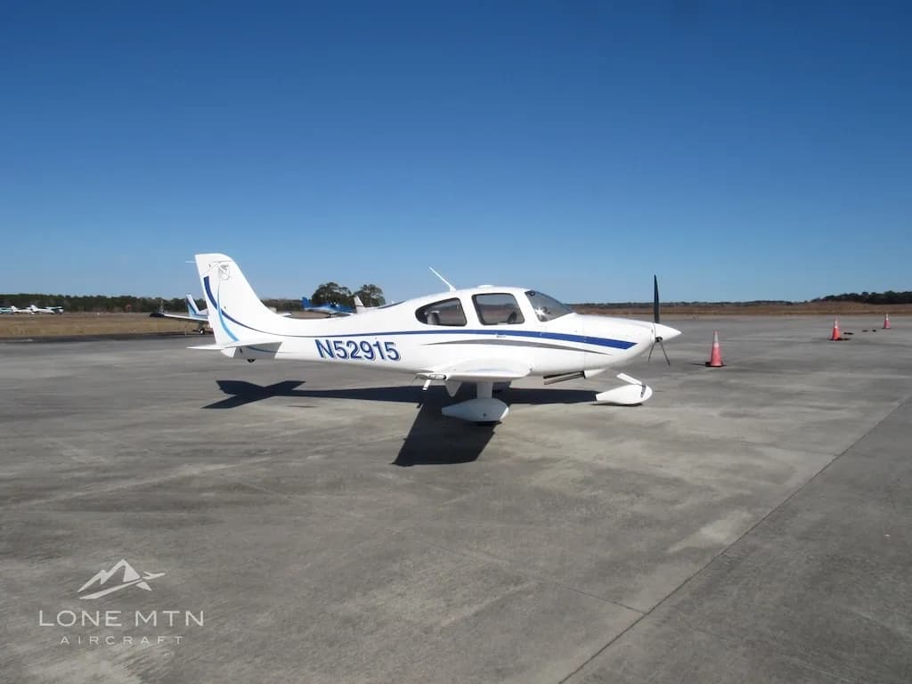 A white Cirrus SR20 aircraft with blue and grey stripes is parked on a concrete ramp, viewed from the front-left.