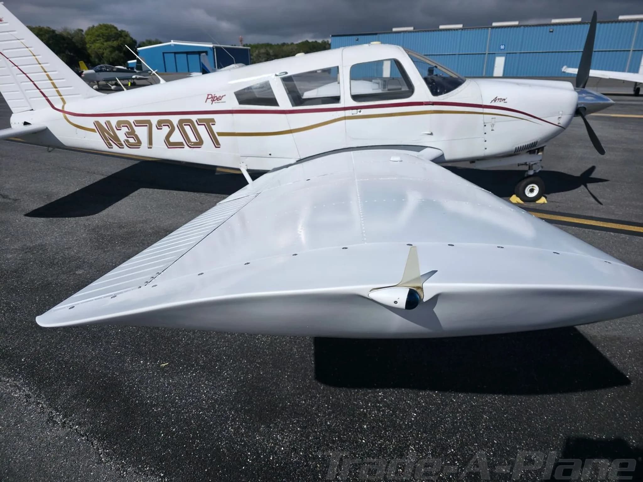 A close-up view of the white Piper Arrow N3720T's wing and fuselage, with blue hangars in the background.