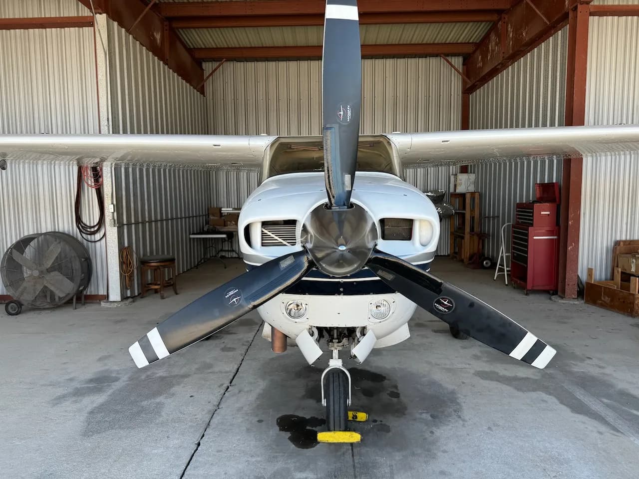 A front view of a white Cessna aircraft with a three-bladed propeller, parked in a hangar.