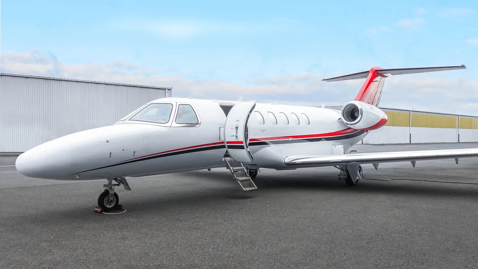A white Citation CJ4 business jet with red and black stripes on the tarmac, with its main cabin door open.