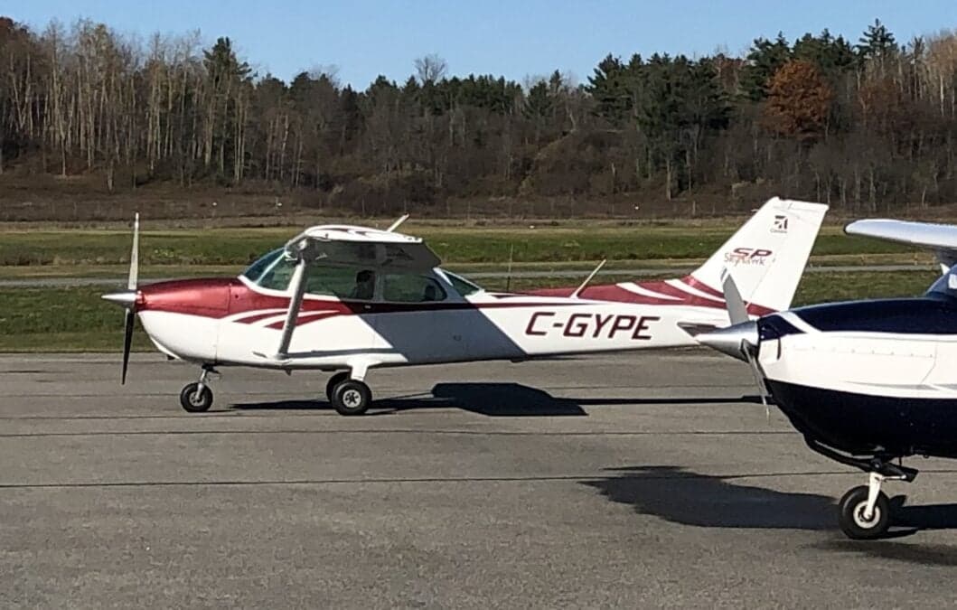 A white and red Cessna 172 (C-GYPE) parked on a tarmac next to another aircraft, with trees in the background.