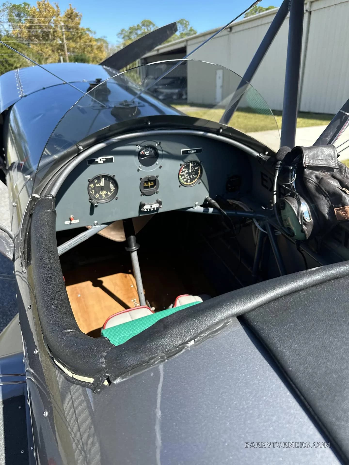 The front cockpit of a gray biplane shows basic flight instruments and a leather headset.