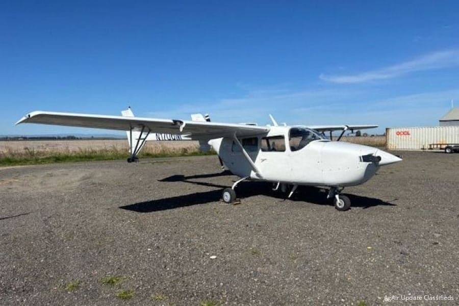 A white, high-wing utility aircraft is parked on a gravel surface under a clear blue sky, with fields and a shipping container in the background.