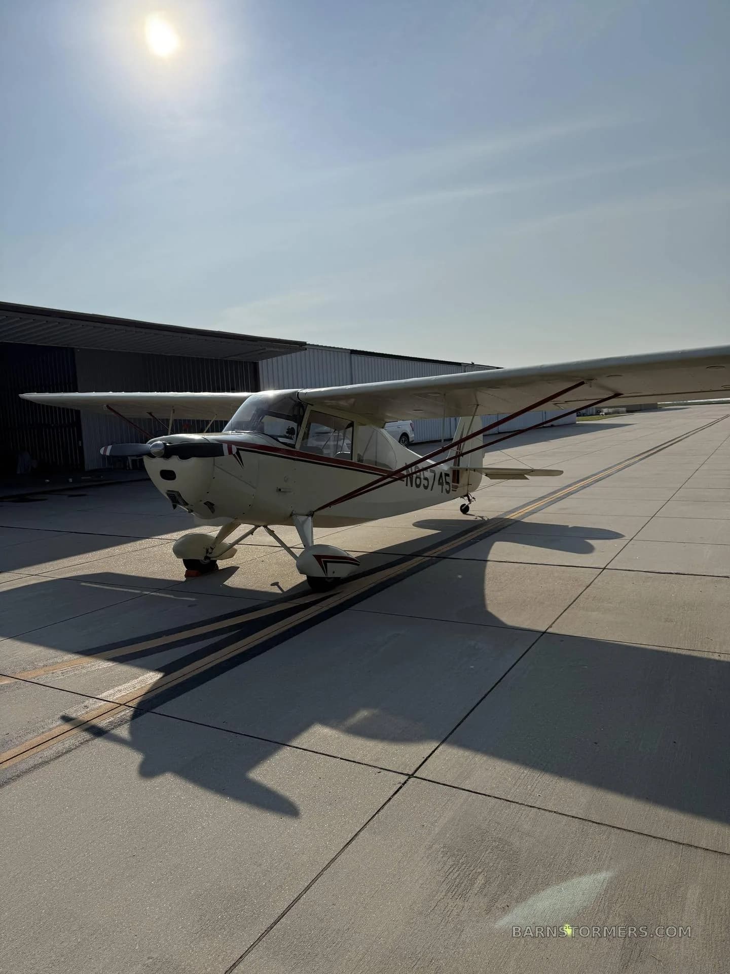 A cream-colored, high-wing taildragger aircraft with red and black stripes, registration N85745, parked on a tarmac with a hangar behind it.