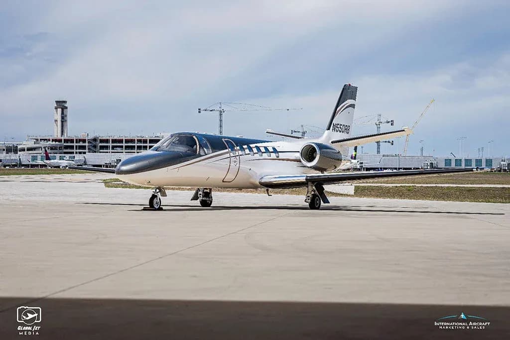 A white and dark grey Cessna Citation V (N550RB) business jet is parked on an airport tarmac with an air traffic control tower in the background.