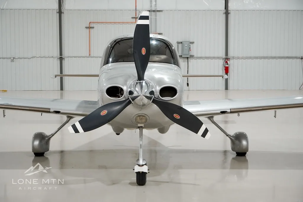 A head-on view of a silver Cirrus aircraft with a three-bladed propeller, parked in a hangar.