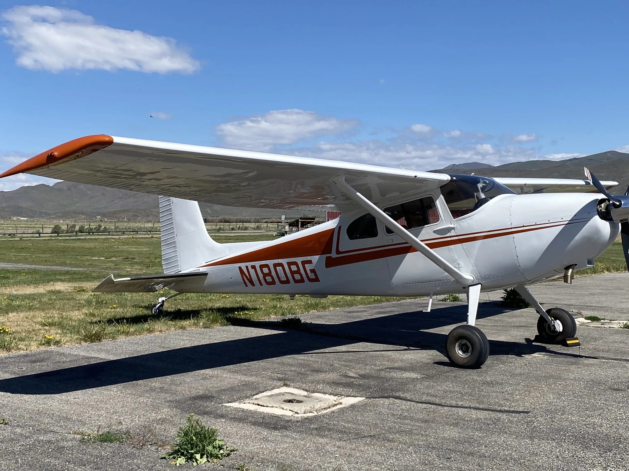 A white Cessna 170B with orange stripes and N180BG registration parked on an airfield with mountains in the background.