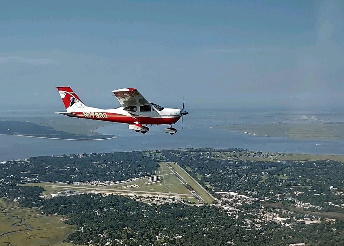 An aerial view of the red and white aircraft flying over a coastal landscape with an airport runway visible below.