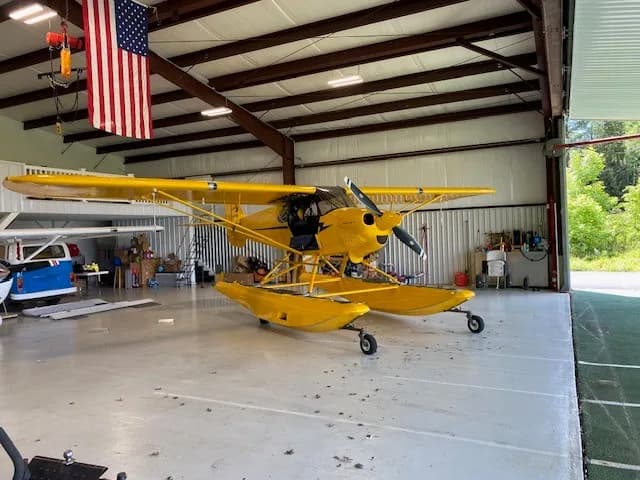 A yellow Carbon Cub floatplane parked inside a hangar with the door open to the outside.