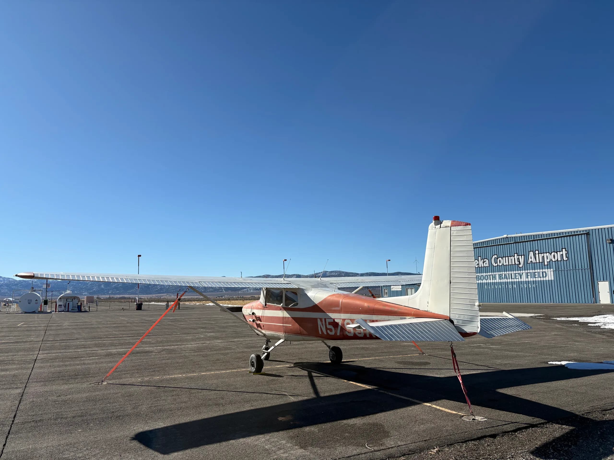 A red and white single-engine high-wing aircraft, N5799A, viewed from the rear-left, parked on an airfield with a blue hangar in the background.