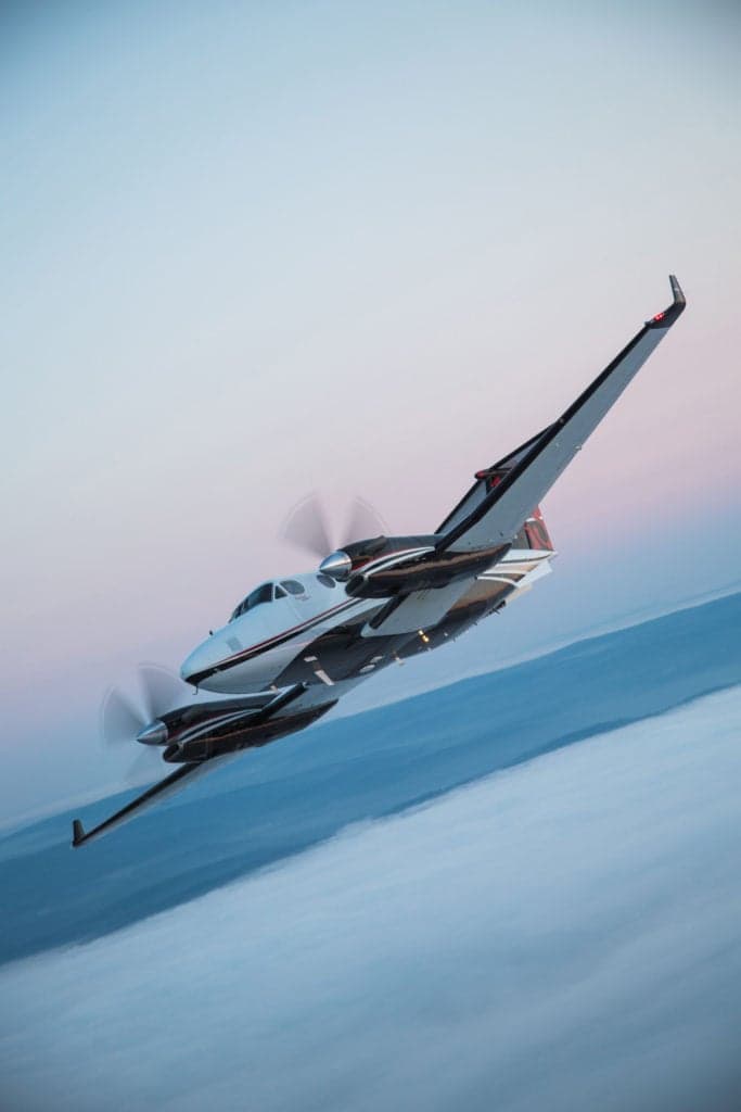 A white and black King Air 350i banks sharply over a cloud layer at dusk.