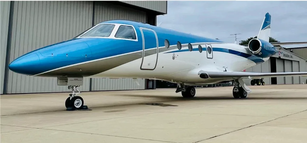 A blue and white Cessna Citation business jet is parked on an airfield next to a hangar.