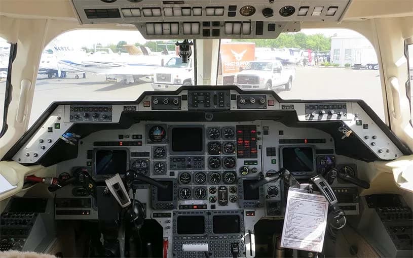 The cockpit of a business jet, showing the instrument panel, yokes, and overhead controls, with other aircraft visible outside.