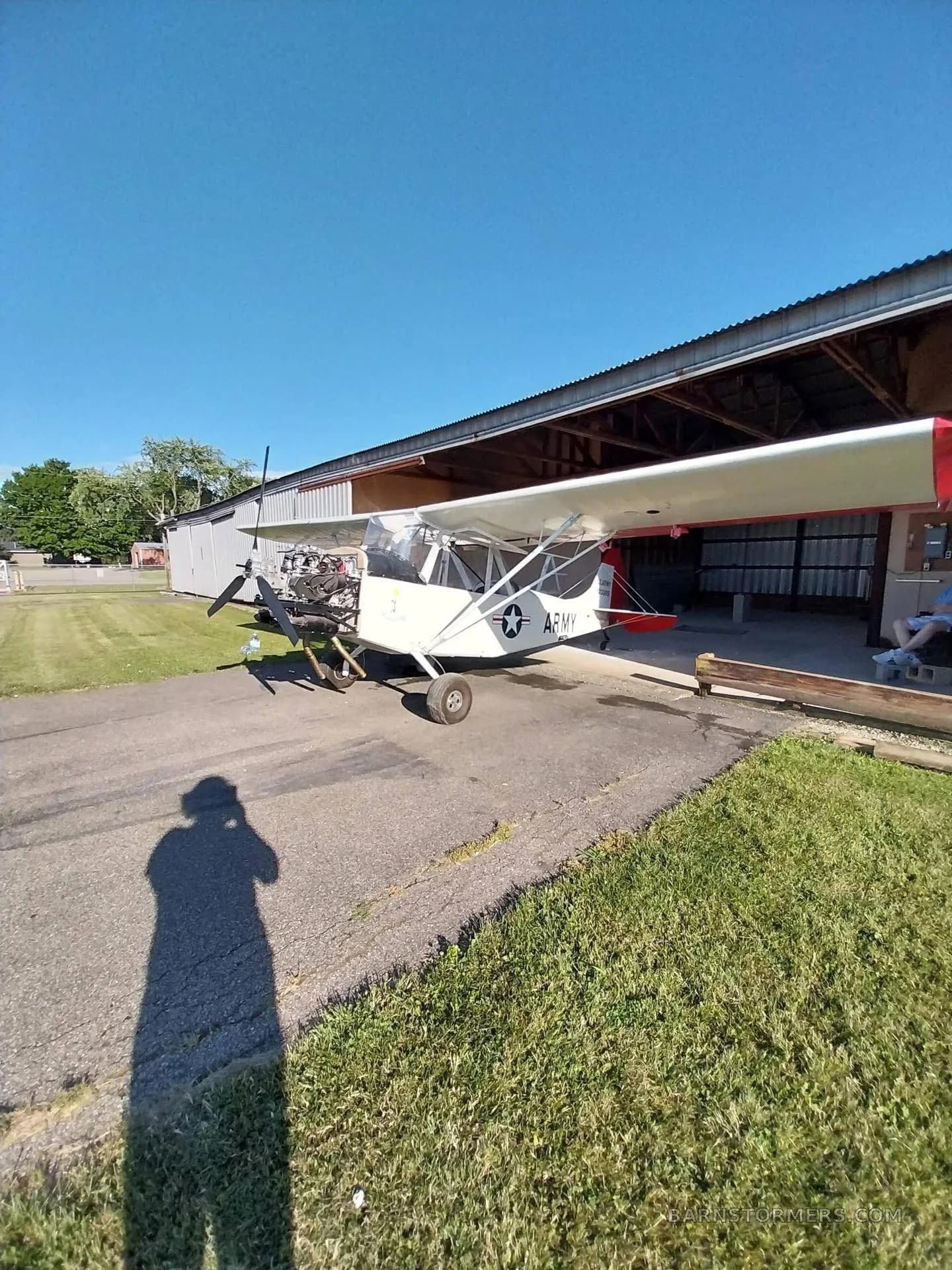 A white experimental aircraft with 'ARMY' markings and an exposed engine is parked on an asphalt ramp outside a hangar on a sunny day.