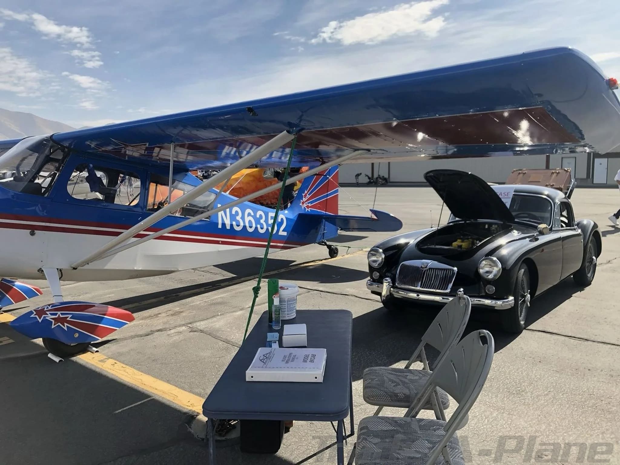 A close-up view of the blue and white high-wing aircraft, N36352, parked on an airfield next to a black classic car with its hood open.
