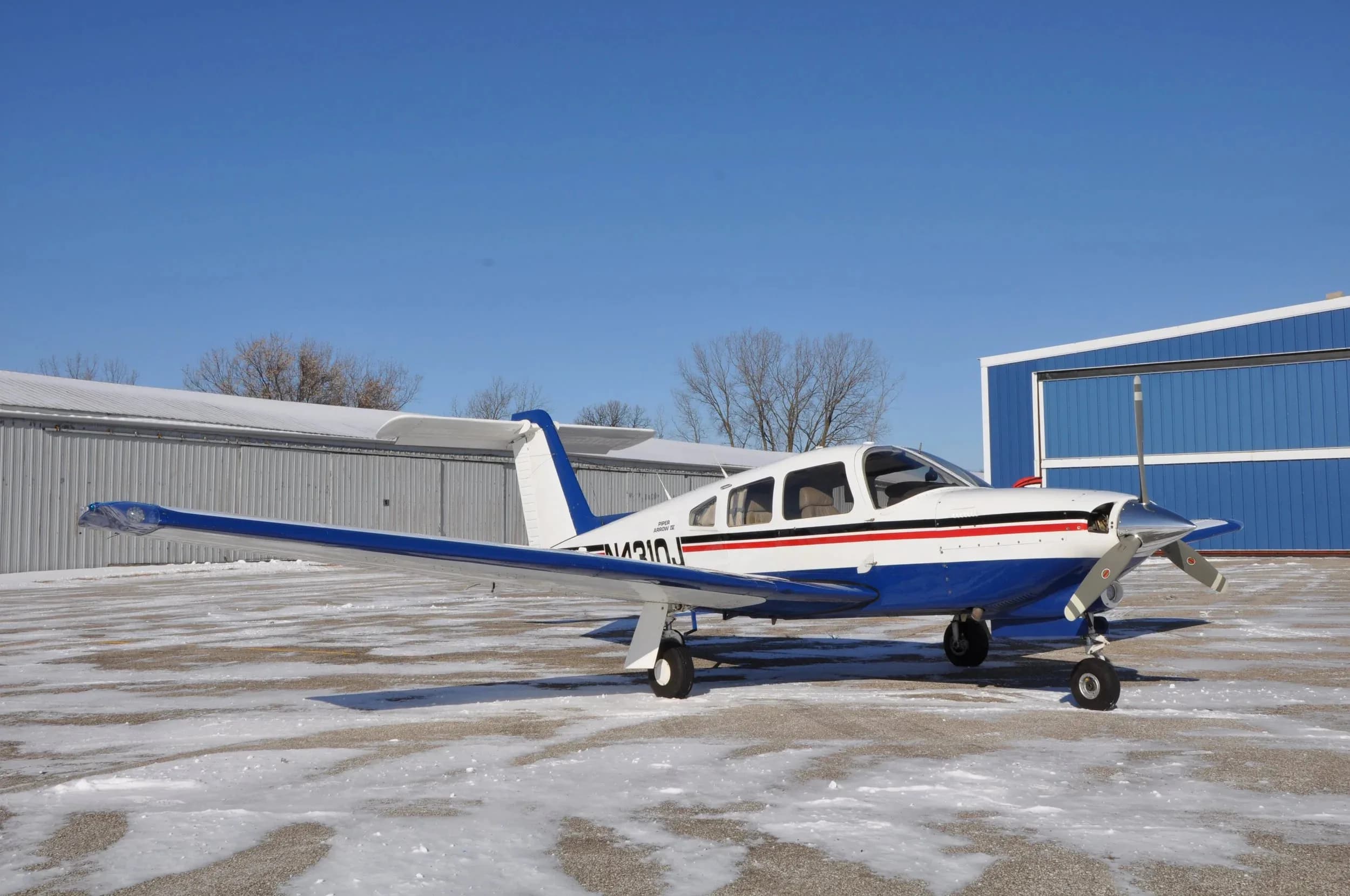 A white and blue Piper Arrow IV aircraft with registration N4310J parked on a snowy tarmac, viewed from the front-left side.