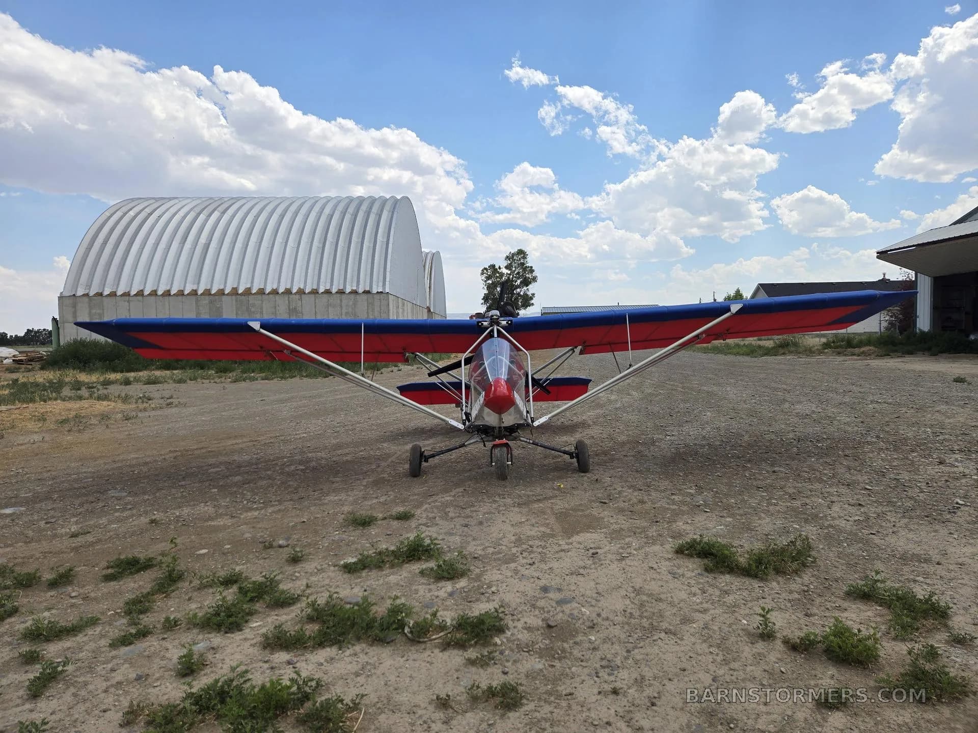 A front view of a red and blue ultralight aircraft with a clear canopy, parked on a dirt field.