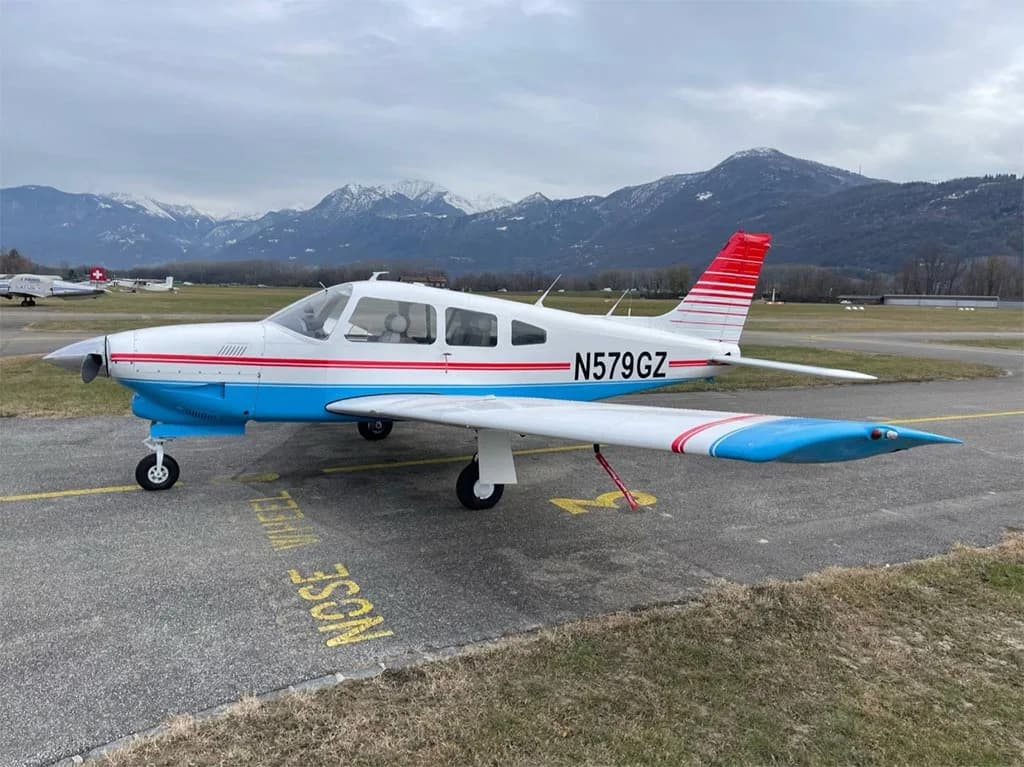 A white and blue Piper PA-28-181 Archer II with red stripes on the tail and wings, parked on an airfield with mountains in the background.