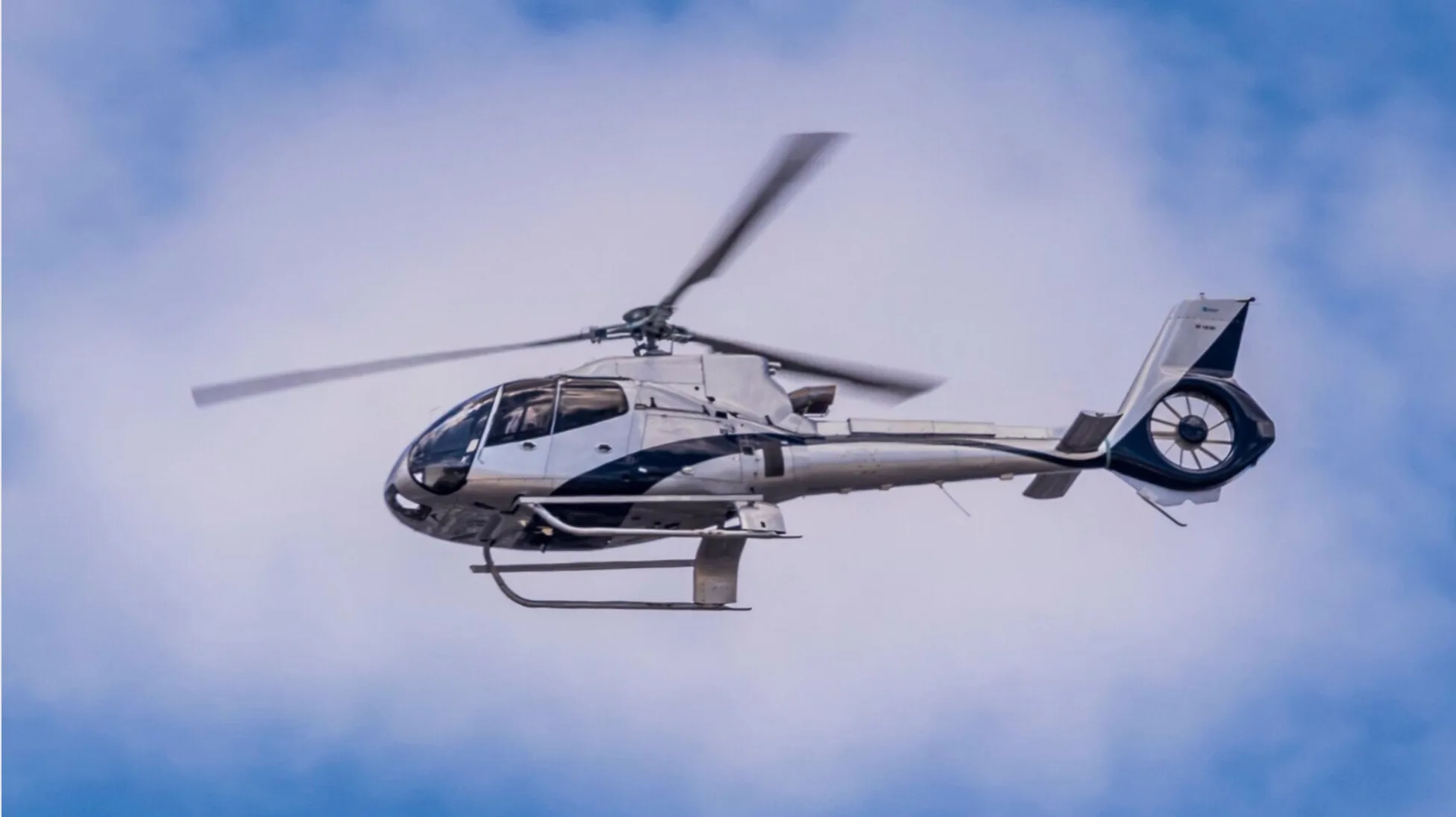 A silver and black Eurocopter EC130 B4 helicopter in flight against a cloudy sky.