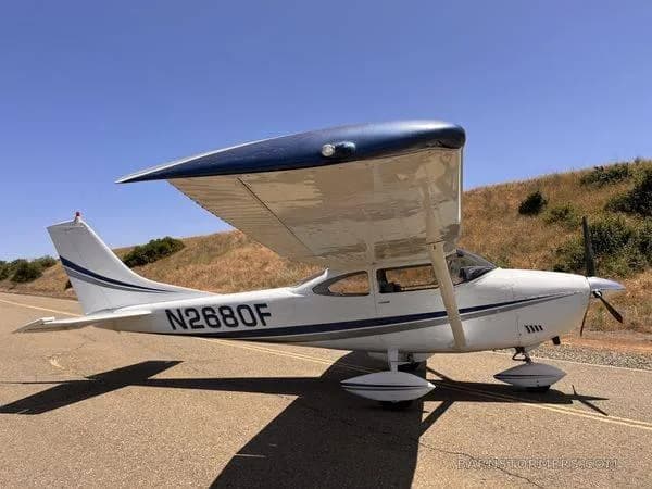 A white single-engine propeller aircraft with blue wing accents and registration N2680F, parked on an asphalt surface under a clear blue sky.