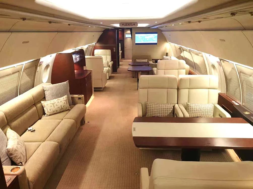 A luxurious private jet cabin with beige sofas, individual seats, and wooden accents, looking towards the rear.