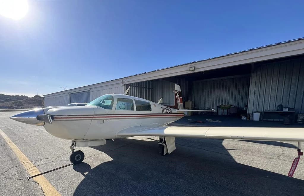 A cream-colored single-engine aircraft with brown and orange stripes, registration N1168B, parked on an asphalt tarmac under a bright sunny sky.