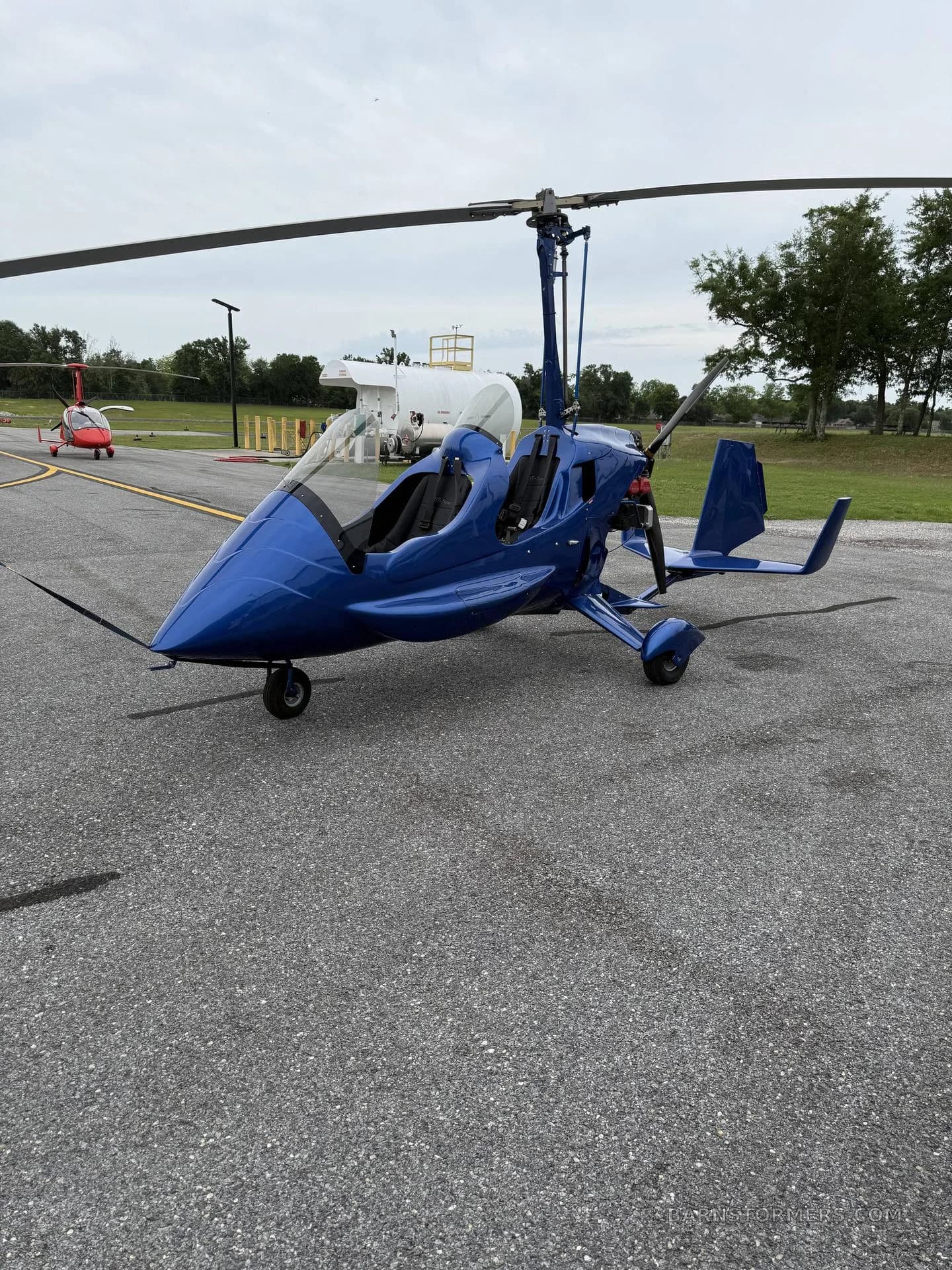 A blue autogyro is parked on an asphalt tarmac, viewed from the front-left, with other aircraft in the distant background.