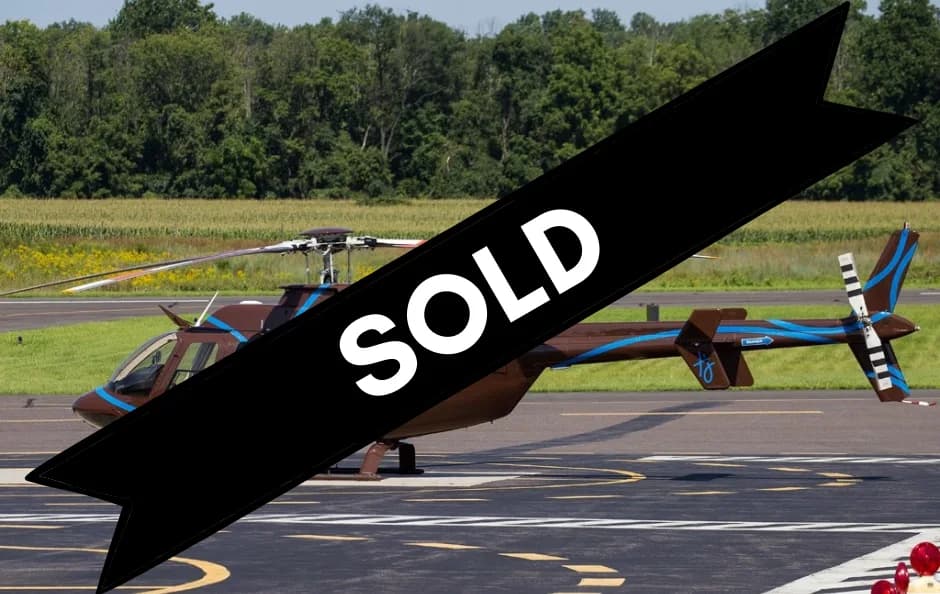 A brown helicopter with blue stripe accents is parked on an airfield, with trees and fields in the background.