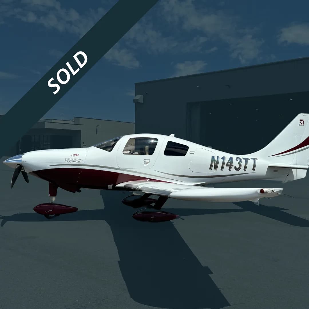 A white and maroon Cessna Corvalis N143TT parked on an asphalt ramp in front of a hangar.