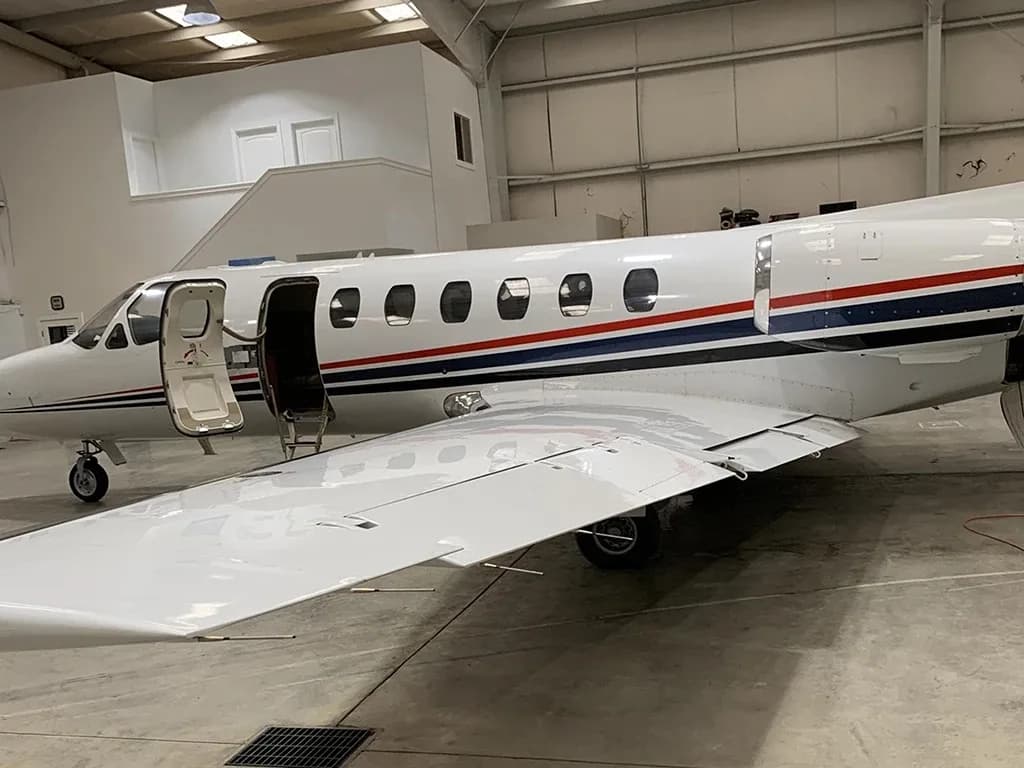 A close-up side view of a white business jet with red and blue stripes, showing the open main door and wing inside a hangar.