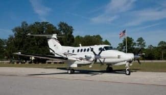 A white and black King Air turboprop aircraft parked on the tarmac with an American flag in the background.