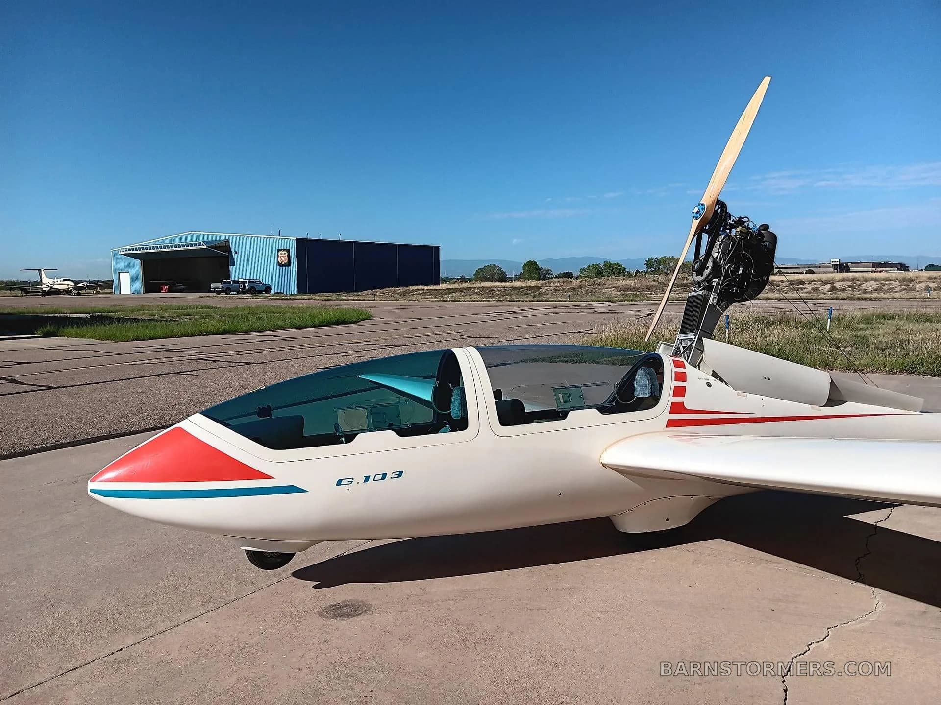A white Grob G 103 glider with its retractable engine and wooden propeller deployed, parked on an airfield tarmac.