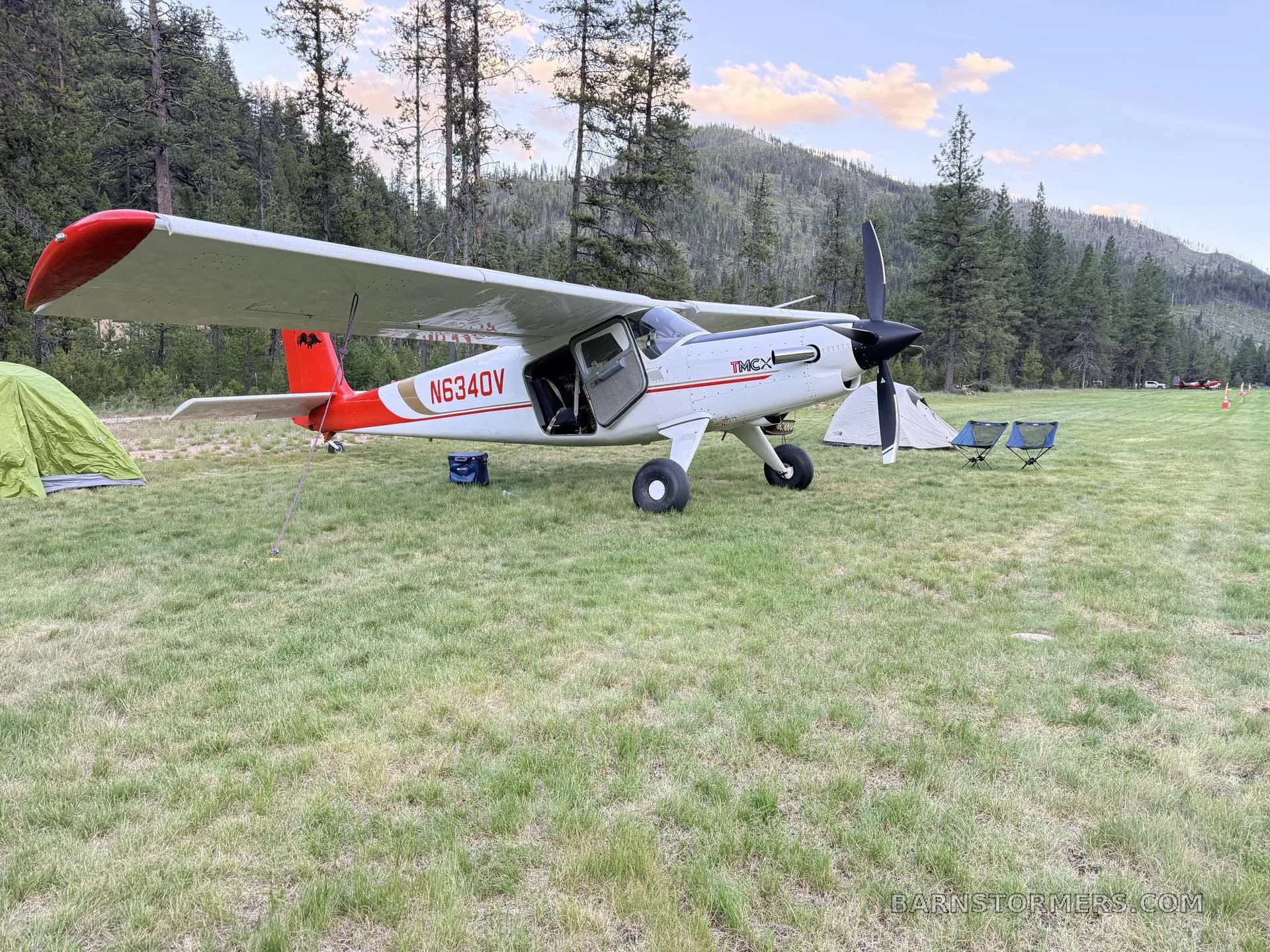 A white and red T-MCX aircraft (N634DV) is parked on a grassy field next to tents and camping chairs, with mountains and trees in the background.