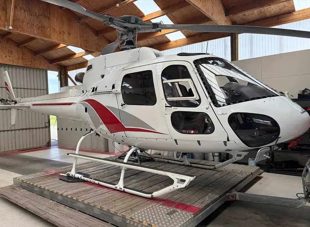 A white helicopter with red and grey stripes, parked on a wooden platform inside a hangar, viewed from the front-right.
