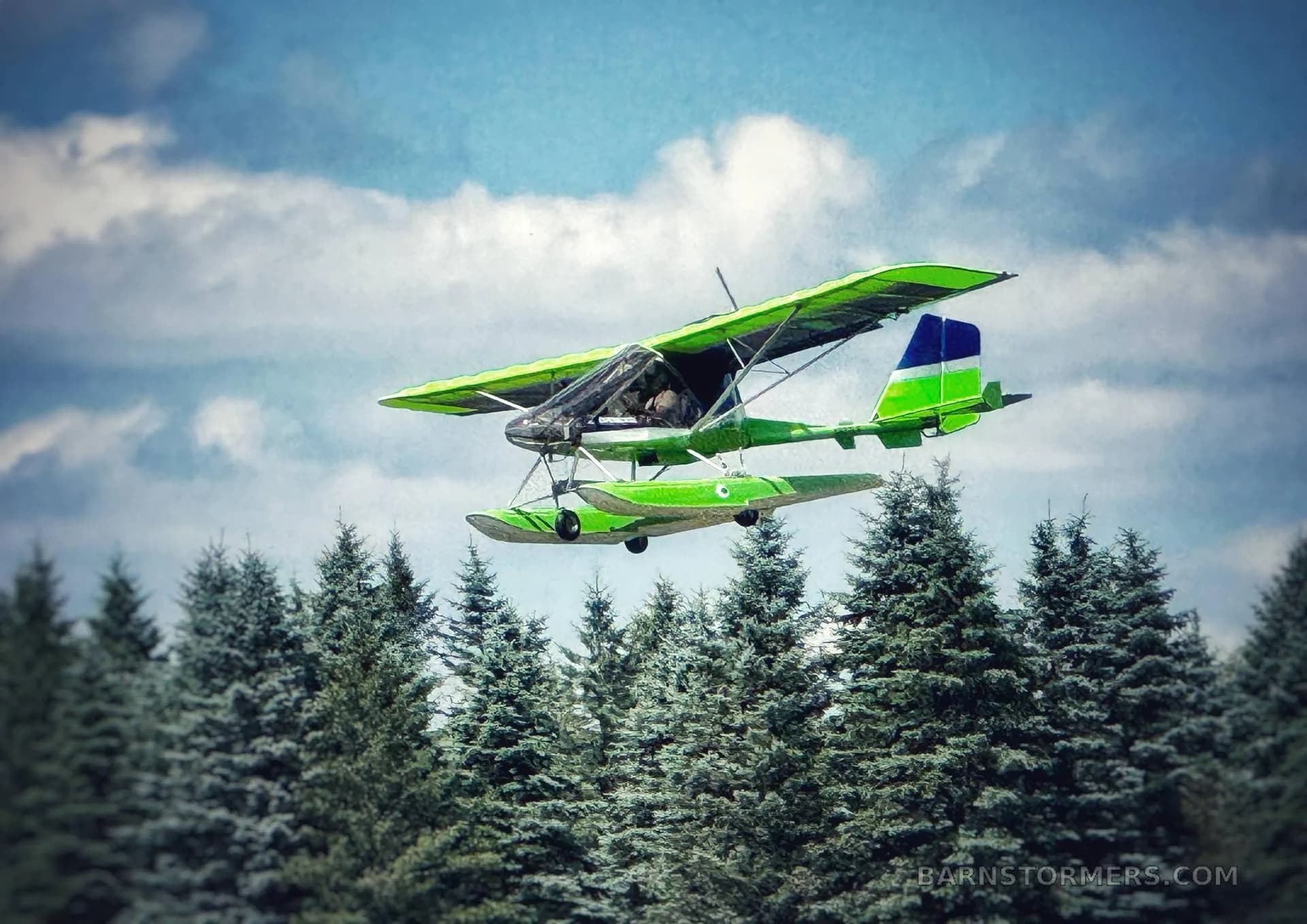 A bright green and blue ultralight seaplane with floats flies low over a line of evergreen trees under a cloudy sky.