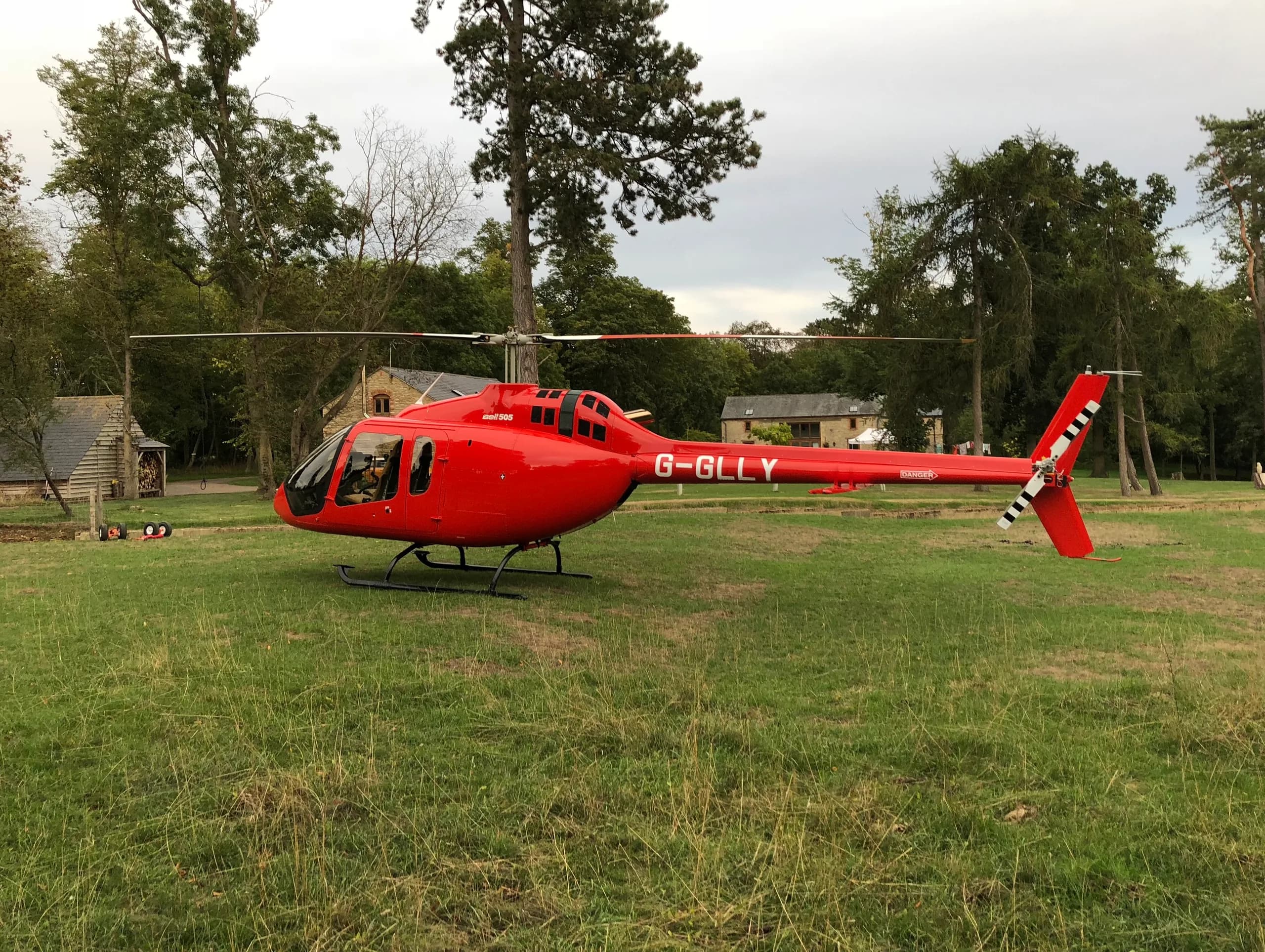 A red Bell 505 helicopter, registration G-GLLY, sits on a grassy field with buildings in the background.