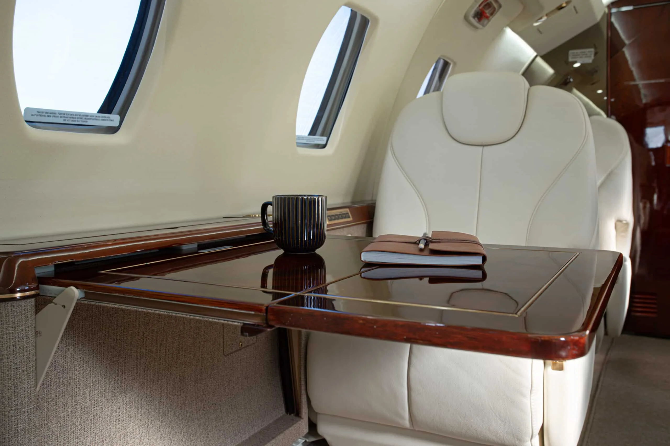 A close-up of a polished wooden table with a black mug and a notebook, next to a cream leather seat in an aircraft cabin.