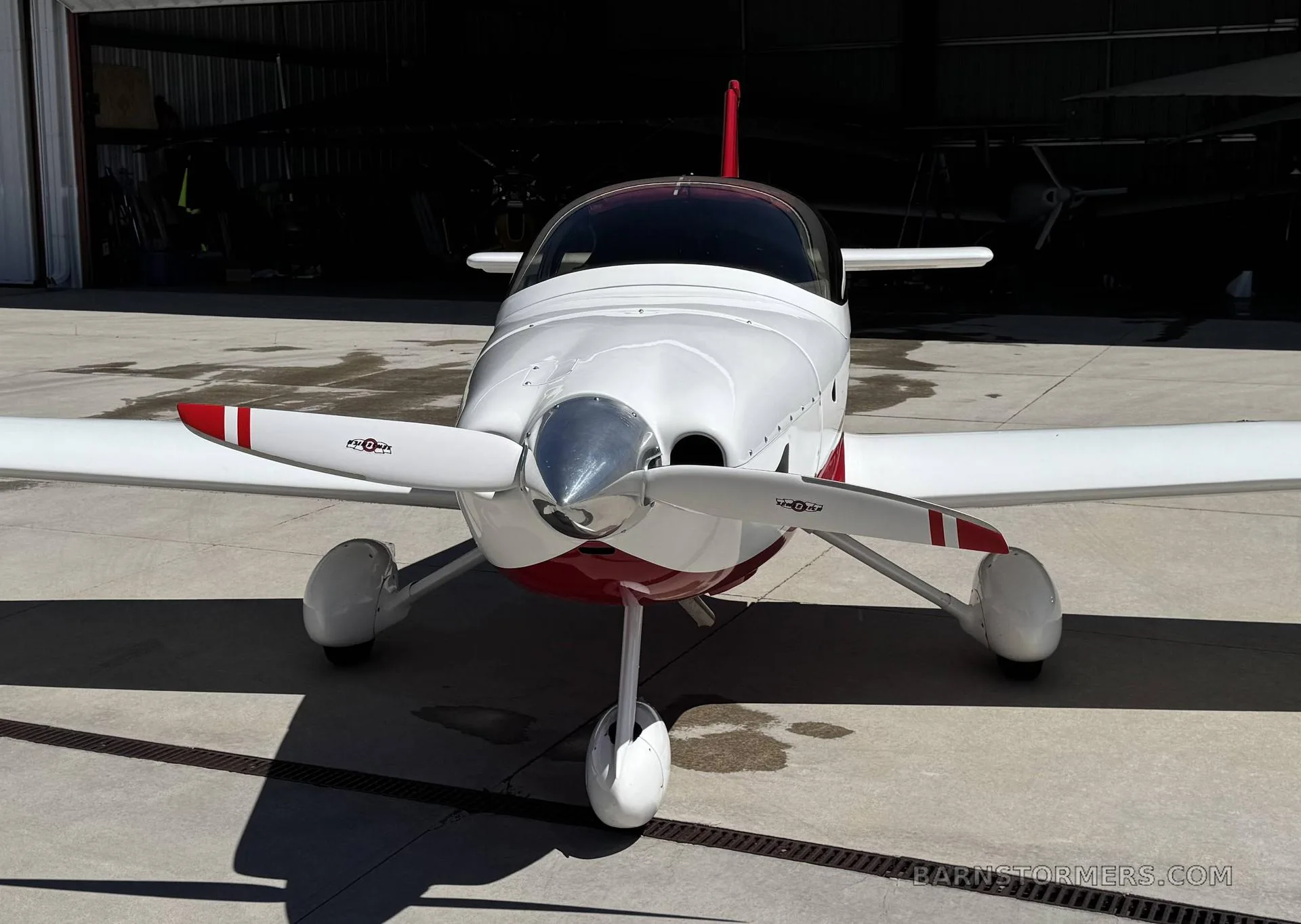 A white and red single-engine aircraft with a propeller, viewed from the front, parked in front of a hangar.